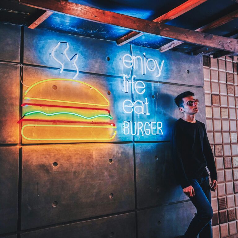 Young man leans against a wall with vibrant neon burger sign in an urban setting.